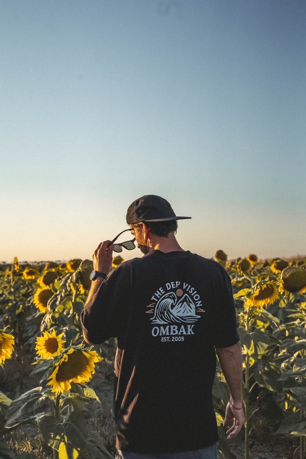 Hombre de espaldas con remera Ombak caminando entre girasoles al atardecer